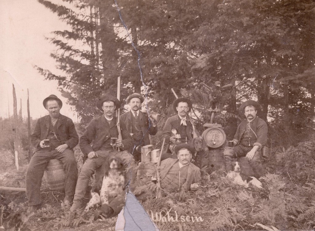 a group of Employees of Doering and Marstrand Brewery in Mount Pleasant pose in the woods holding various firearms and mugs of beer, with a cask of craft beer and several hunting dogs.