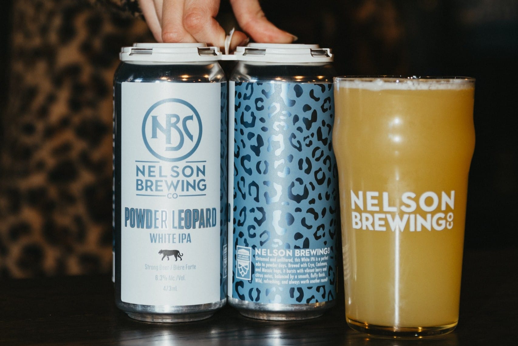 a glass of Powder Leopard White IPA next to a four pack of cans. Brewed by Nelson Brewing Company